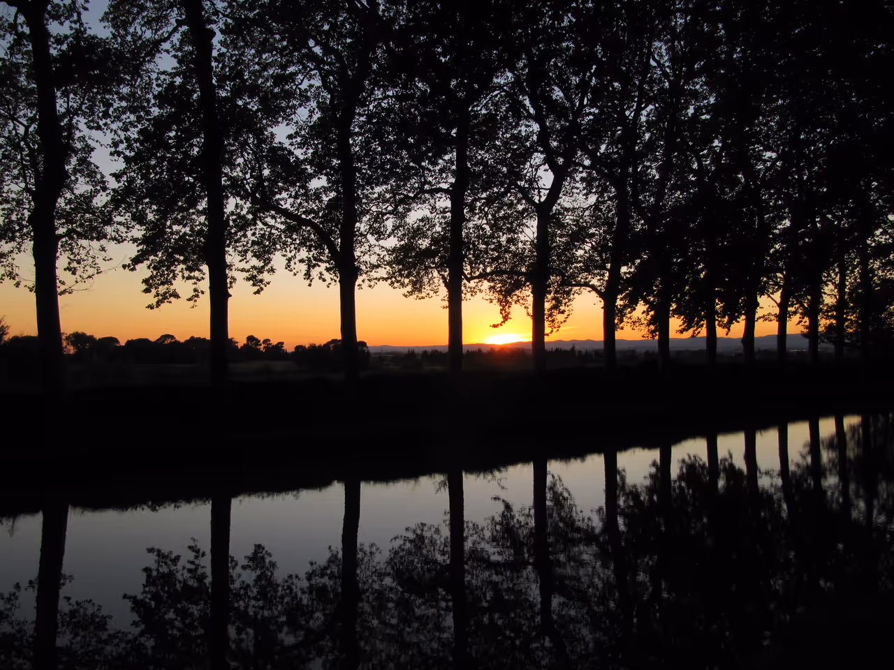 Sunset over the Canal du Midi near Colombiers with trees silhouetted against an orange sky and reflecting in the water