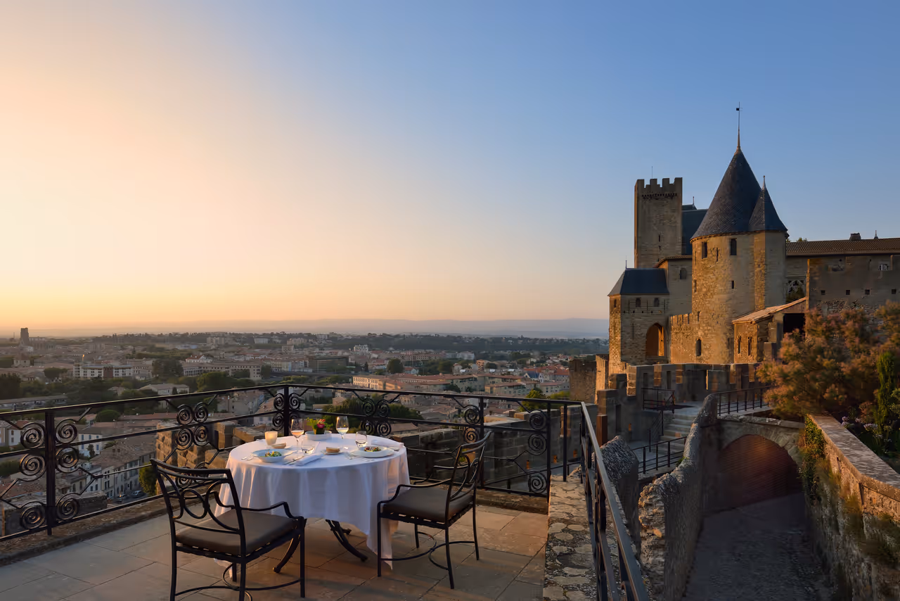 Panoramic terrace of La Barbacane restaurant with a set table overlooking the medieval ramparts of Carcassonne and the lower town at sunset
