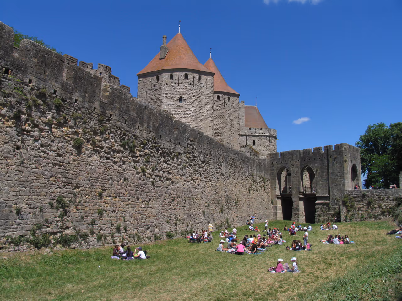 People relaxing on the grass inside the medieval fortified walls of the Cité de Carcassonne under a clear blue sky