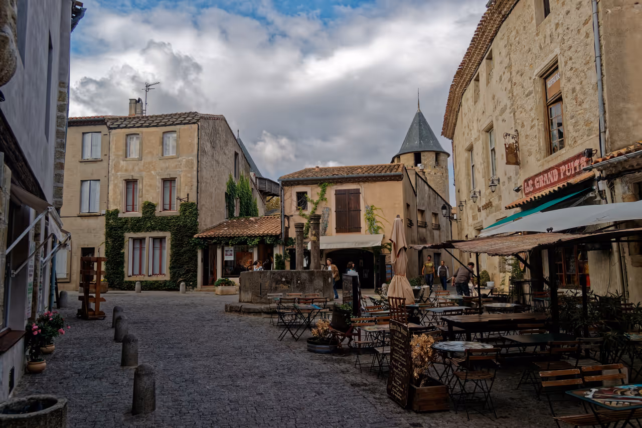 Cobblestone square at Place du Grand Puits in the medieval fortified city of Carcassonne with outdoor restaurant seating, stone buildings and a conical tower under a cloudy sky