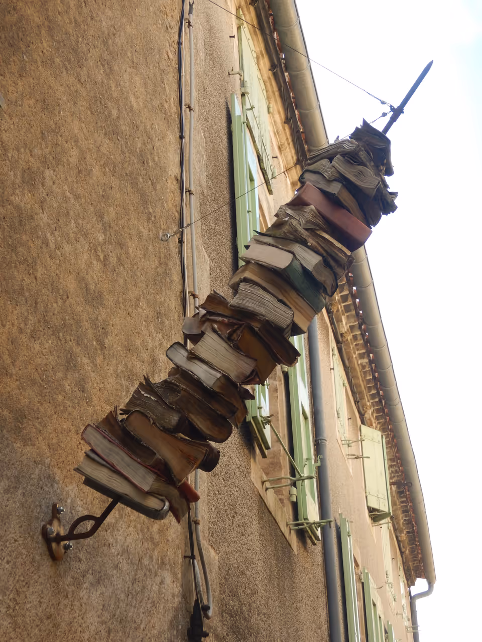 Outdoor sculpture of books stacked on a metal spit against a stone building in Montolieu Village du Livre