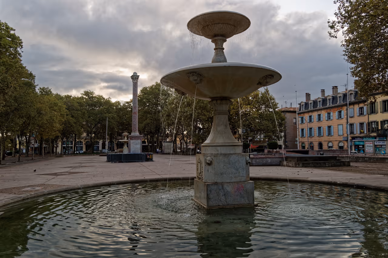 Water fountains in Jardin de Andre Chenier with a column and buildings in the background under a cloudy sky in Carcassonne