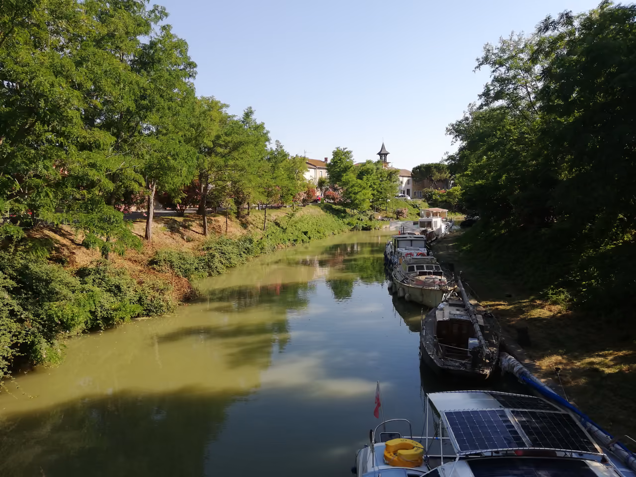 Boats moored on the calm waters of the Canal du Midi in Poilhes surrounded by lush green trees and village buildings