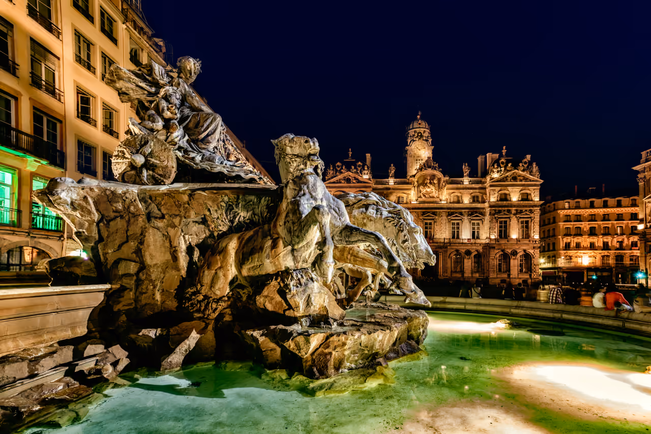 Night view of the Bartholdi Fountain at Place des Terreaux in Lyon featuring the illuminated sculpture of a woman steering a chariot pulled by four horses