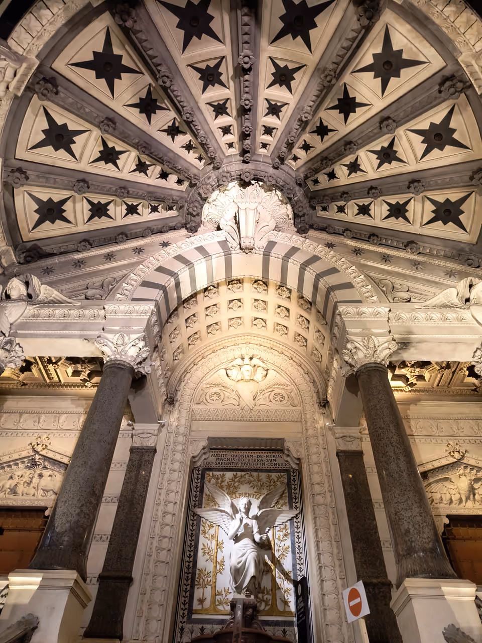 Ornate vaulted ceiling with geometric star patterns above an angel statue inside the Basilica of Notre-Dame de Fourviere
