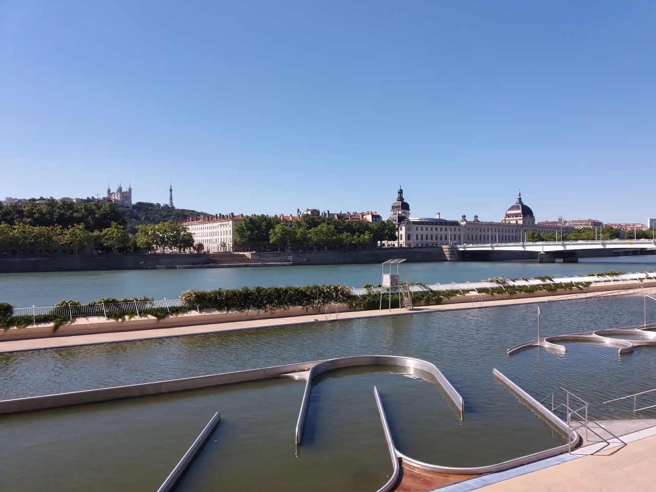 An outdoor swimming pool complex along the Rhône river in Lyon with the city skyline and bridges in the background