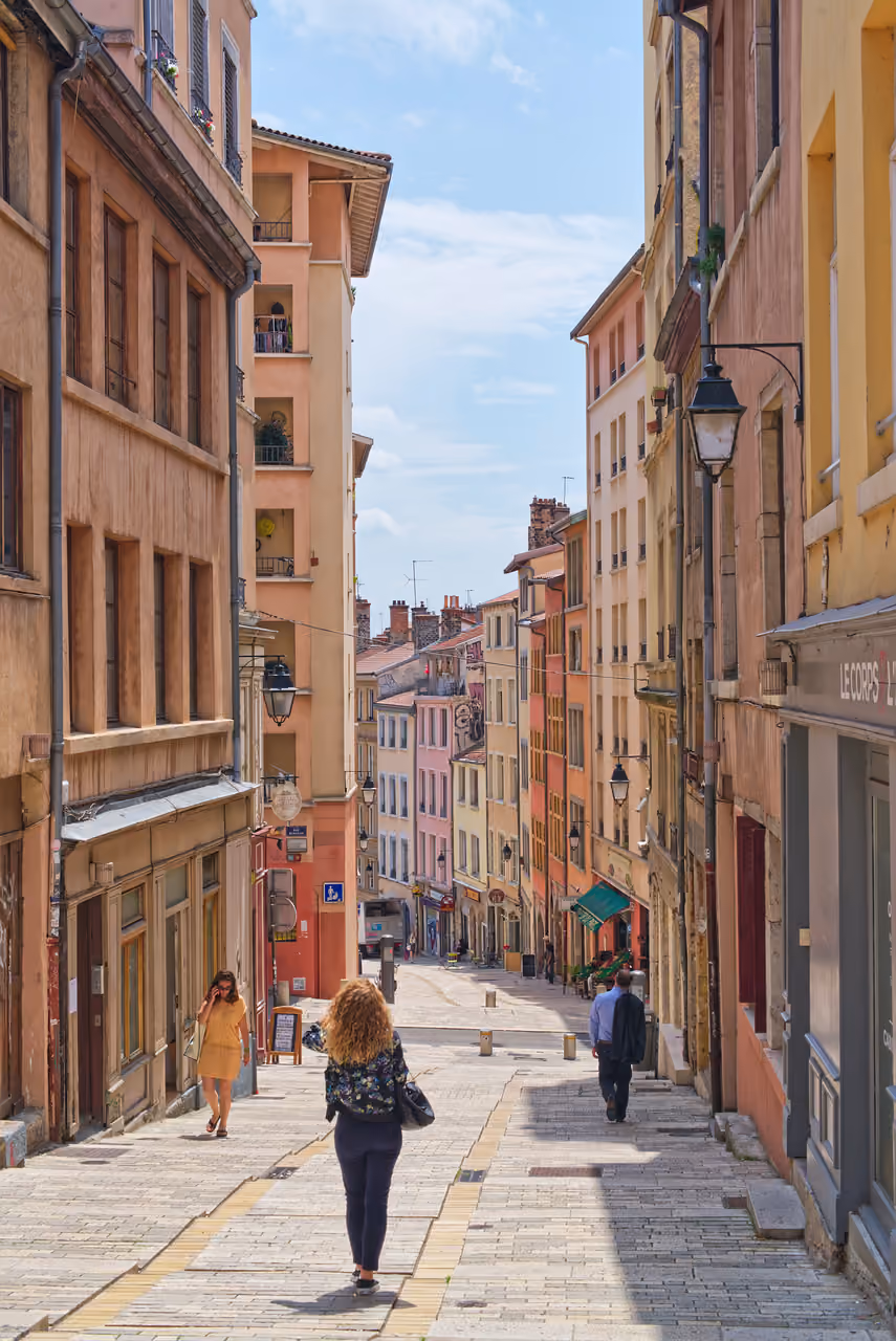 A scenic pedestrian street with historic multi-storey buildings in Lyon, France, featuring people walking on a sunny day