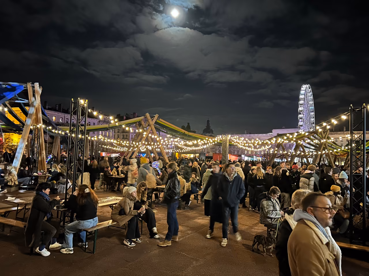 Crowds enjoying the Fête des Lumières festival at night with market stalls, overhead string lights and a illuminated Ferris wheel in Place Bellecour