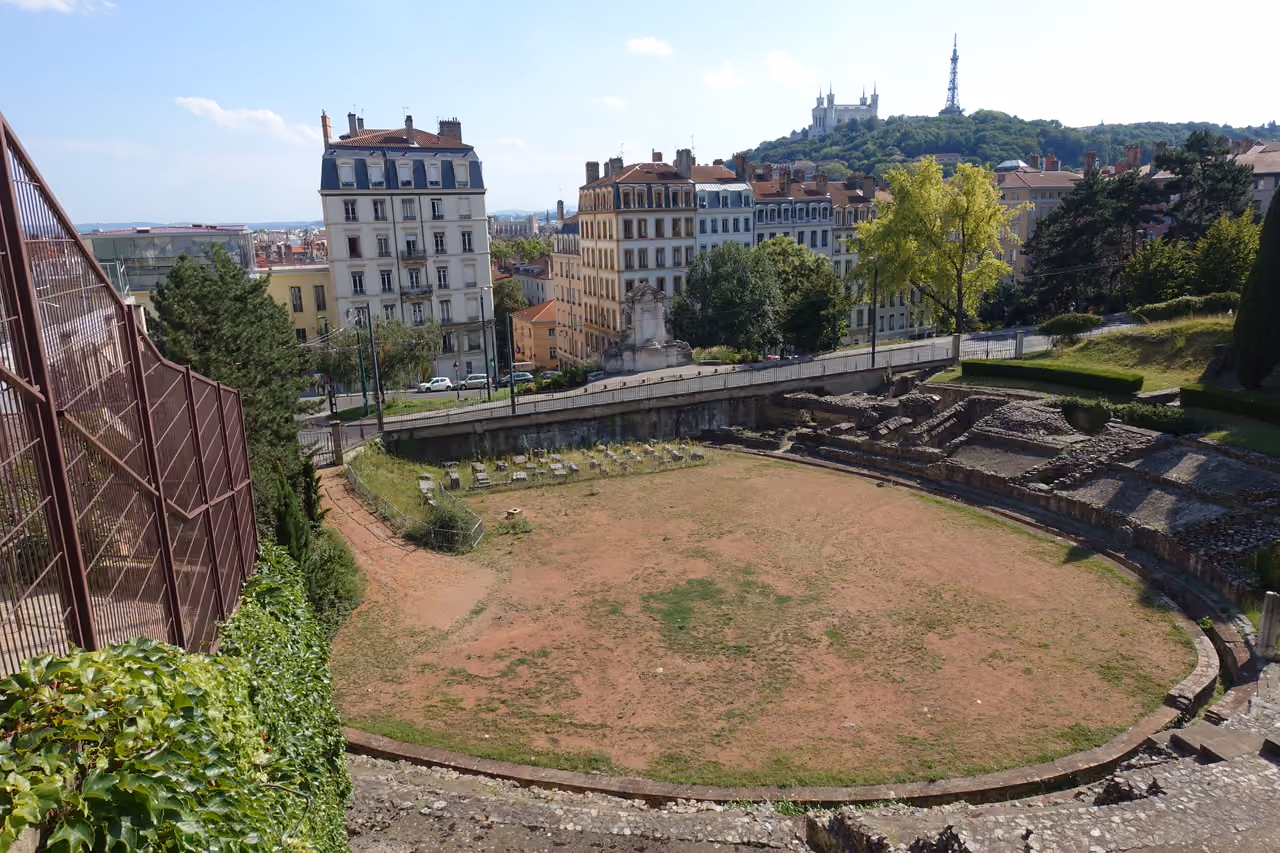 An elevated view of the ancient Roman Amphitheatre of the Three Gauls in the Croix-Rousse district, showing the semicircular stone ruins and arena floor against a backdrop of historic city buildings and a hilltop church