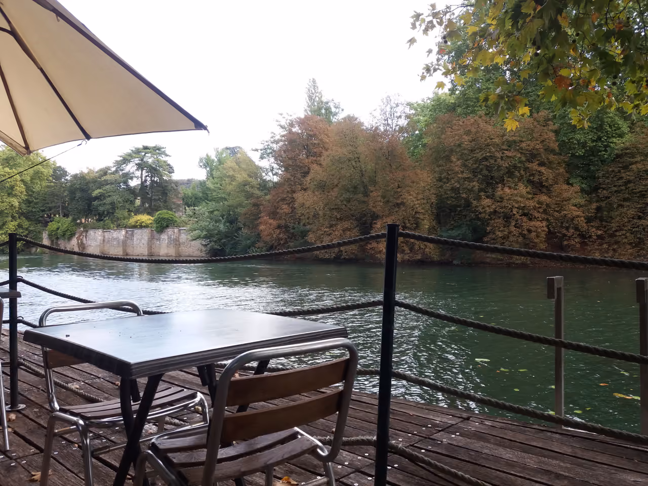 Outdoor restaurant terrace with wooden tables and chairs overlooking a scenic river and lush green trees