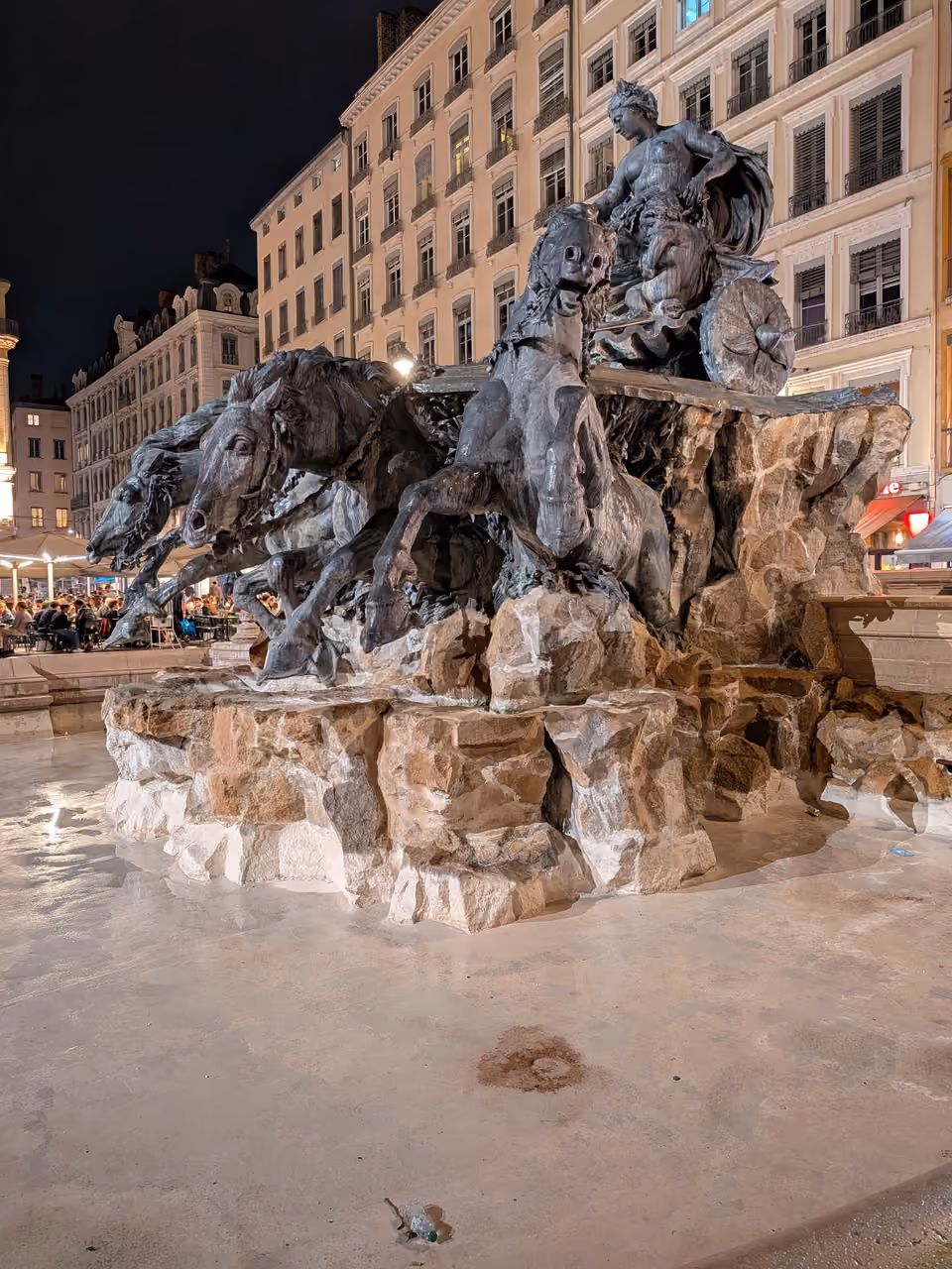 The Bartholdi Fountain lit up at night on Place des Terreaux featuring a dramatic sculpture of a woman steering a chariot pulled by four rearing horses