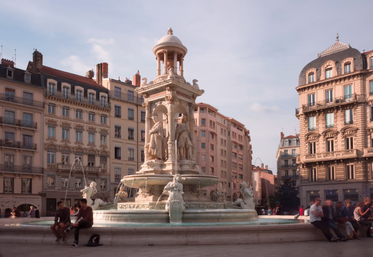 The Fontaine des Jacobins in Lyon features an ornate white marble fountain surrounded by historic European architecture under a clear blue sky
