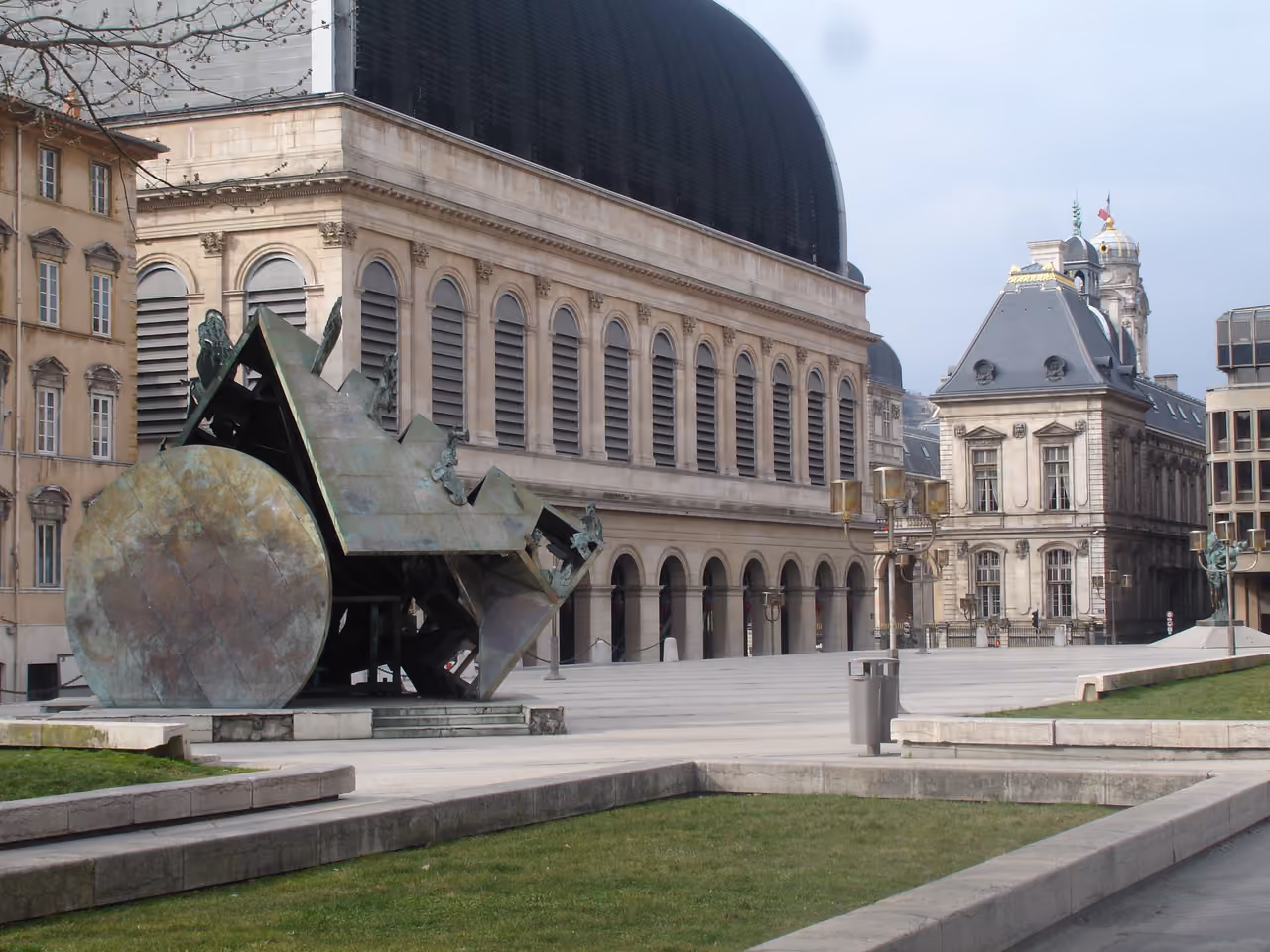 Modern public square featuring a large abstract bronze fountain sculpture in front of an opera house with a arched vaulted roof