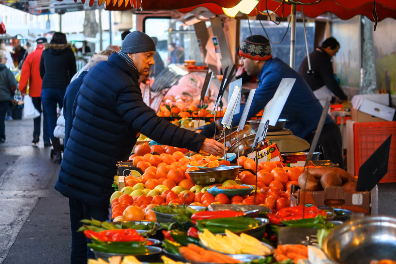 A shopper browsing fresh oranges, peppers, and vegetables at an outdoor market stall