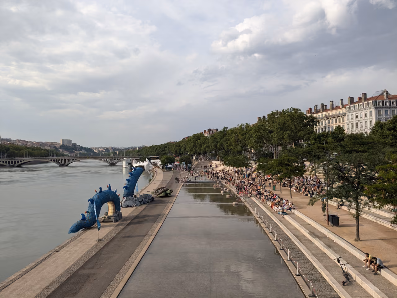 Crowds gathered at the waterfront promenade of Berge Marie-Sklodowska-Curie featuring a blue sea serpent sculpture along the river Rhône