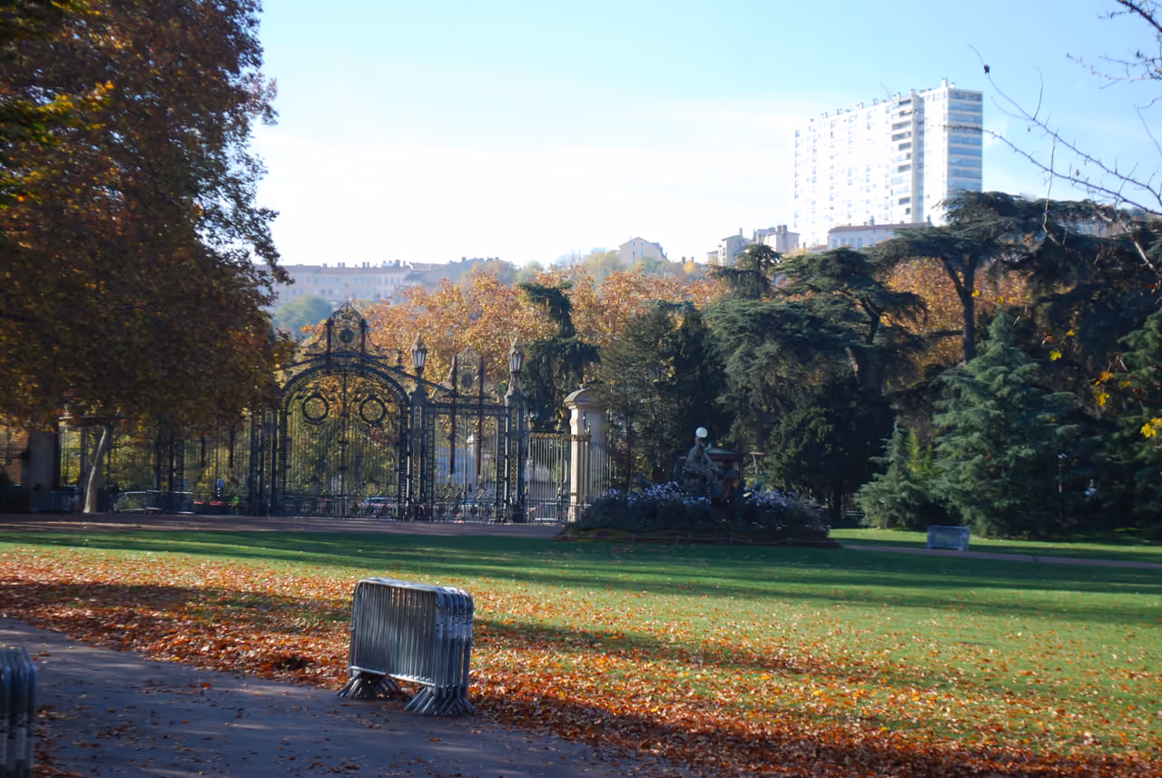 Large ornate iron gates at the entrance of Parc de la Tête d'Or in autumn with fallen leaves on the grass and trees in the background