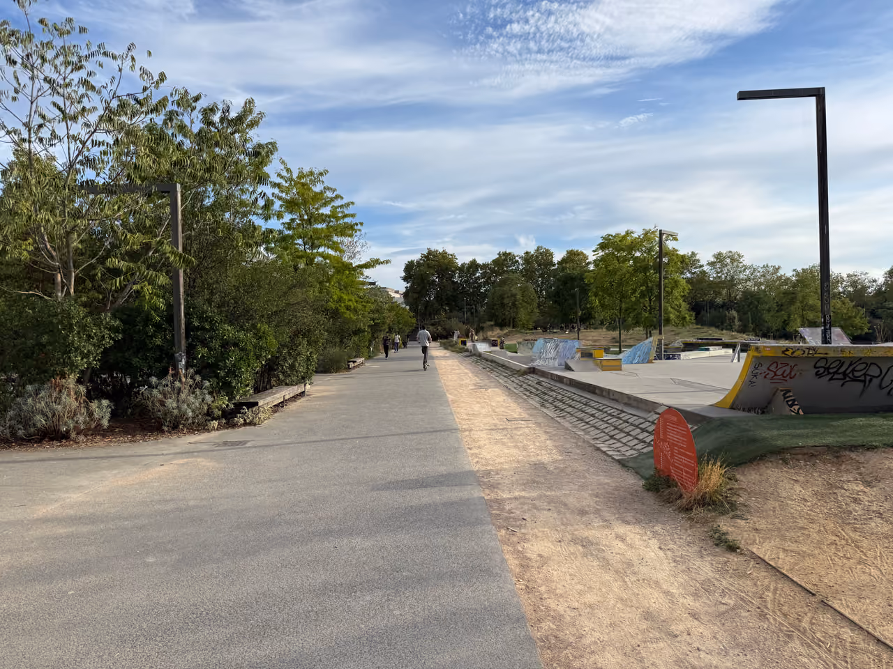 Paved walking path next to a skatepark in Parc Sergent Blandan with trees and a blue sky