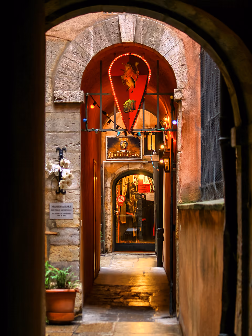 Narrow stone alleyway in a historic traboule with an arched entryway, decorative lights, and signs leading to a medieval-style shop
