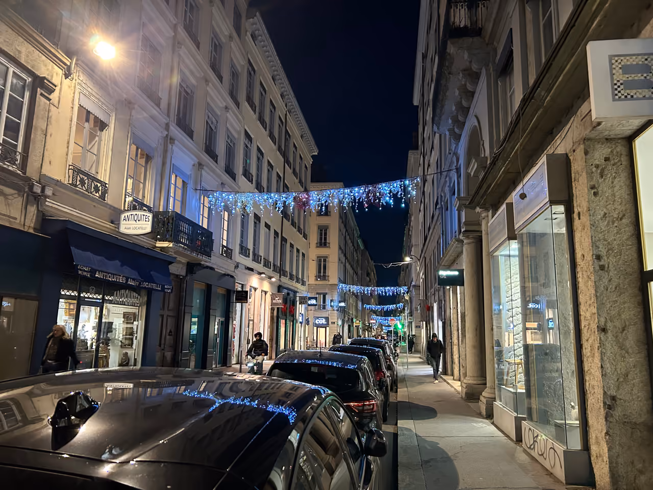 European historic city street at night decorated with festive blue and white Christmas lights stringing across buildings above parked cars