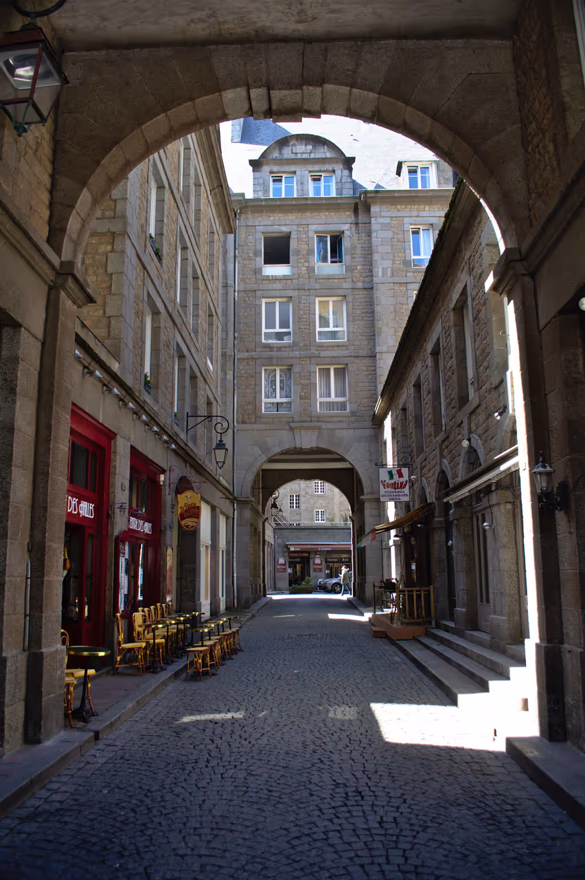 Narrow cobblestone street framed by an archway in the walled historic old town of Saint-Malo with stone buildings and dining tables