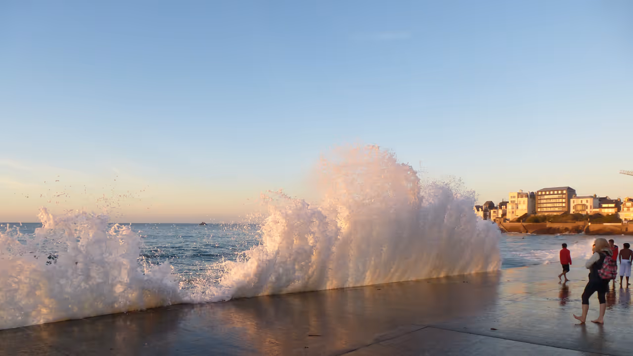 Powerful wave crashing against a seawall at sunset, sending a spray of water into the air as beachgoers watch from the shore, Saint-malo