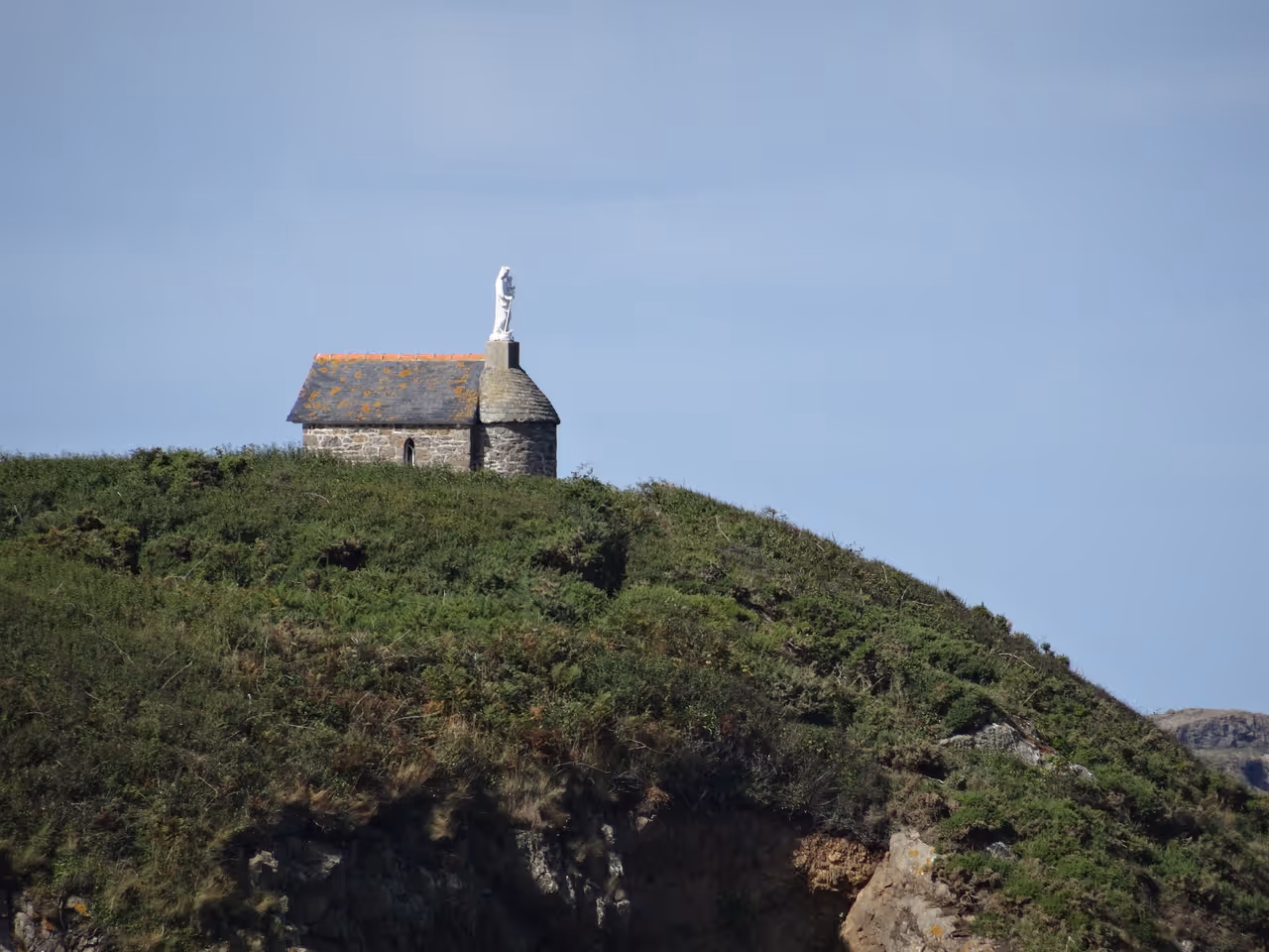 Stone chapel built on a grassy clifftop with a white statue of the Virgin Mary rising above the roof overlooking the Emerald Coast near Saint-Malo