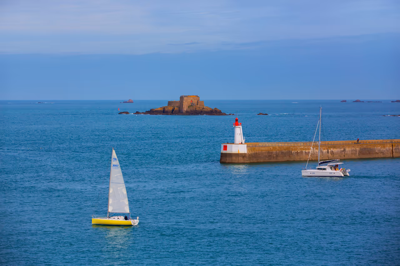 Sailing boat and catamaran on the emerald sea near a white lighthouse pier and a historic tidal fort in Saint-Malo