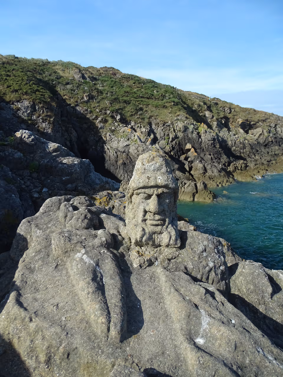 Sculpted granite faces of Rochers sculptés de Rothéneuf carved into the coastal rocks along the blue ocean water in Brittany