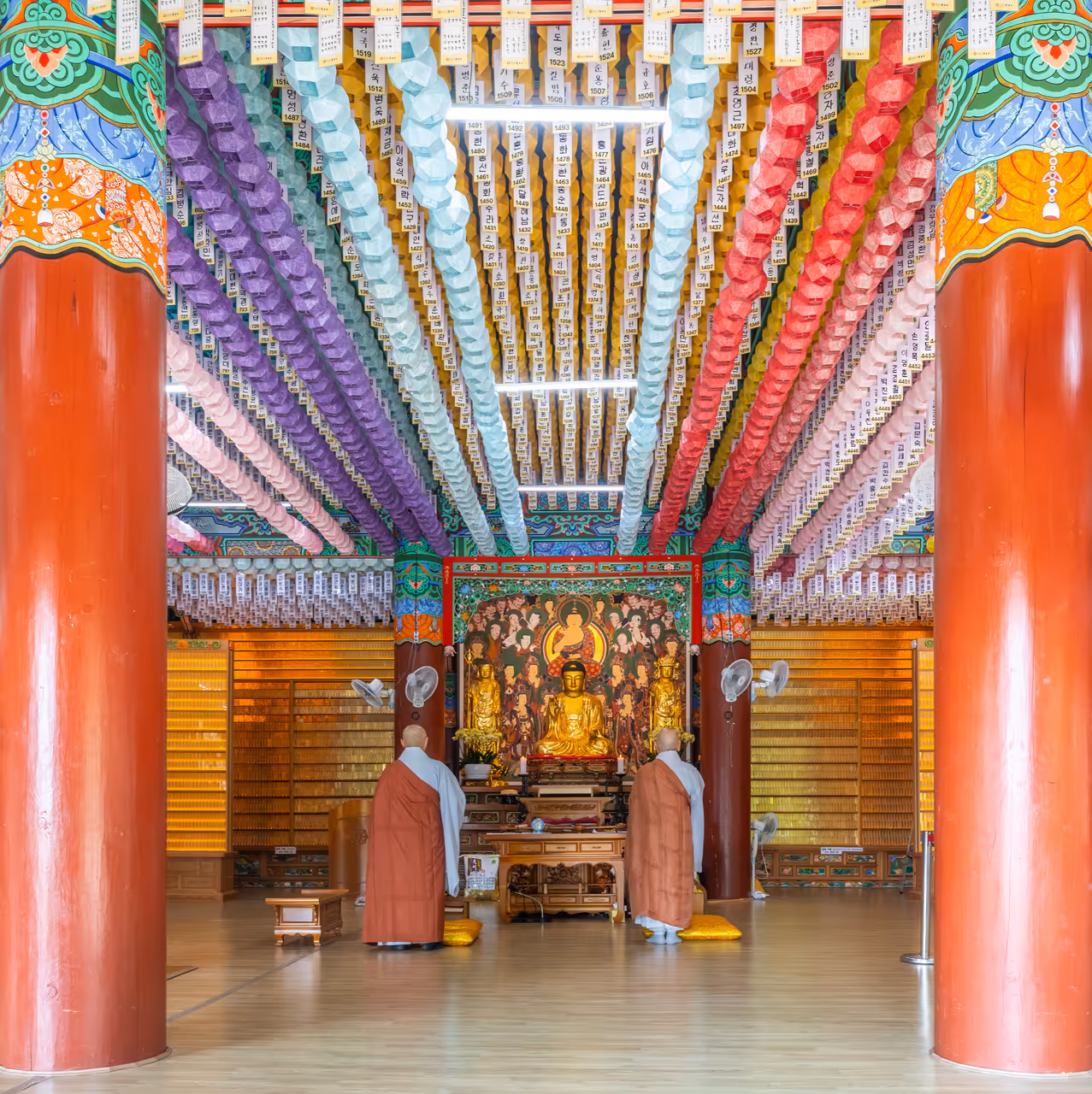 Interior view of Beomeosa Buddhist temple during a ceremony, with paper lanterns on the ceiling, a gold statue of the seated Buddha, and 2 Buddhist monks, in Busan