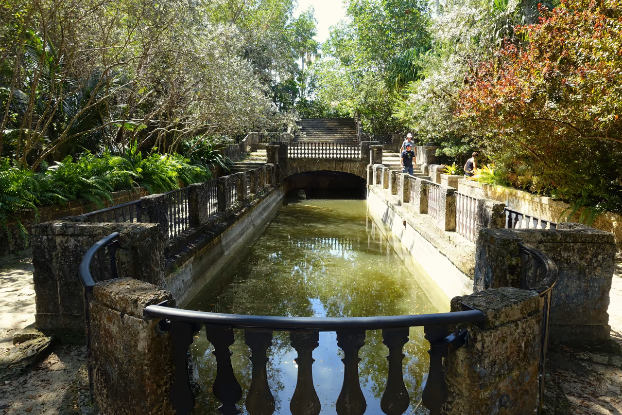 An ornate stone swimming pool canal surrounded by lush gardens, ferns, and mature trees