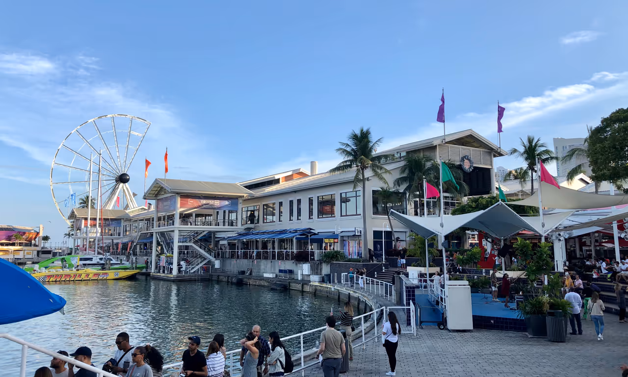 A sunny day at Bayside Marketplace in Miami showing the harbour, palm trees, outdoor dining areas, and the Skyviews Miami Ferris wheel under construction