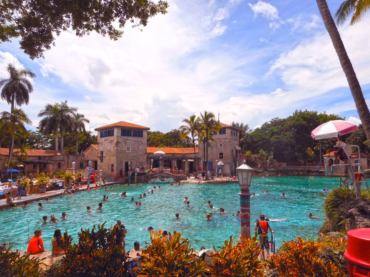 Crowded Venetian Pool in Coral Gables with tourists swimming in clear turquoise water, historic Mediterranean revival buildings and tropical palm trees under a blue sky
