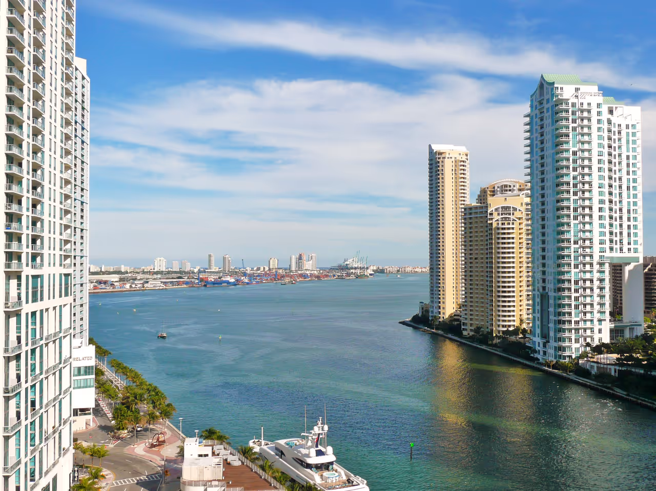 Aerial view of modern waterfront skyscrapers flanking the mouth of the Miami River as it flows into Biscayne Bay near a bustling shipping port under a clear blue sky
