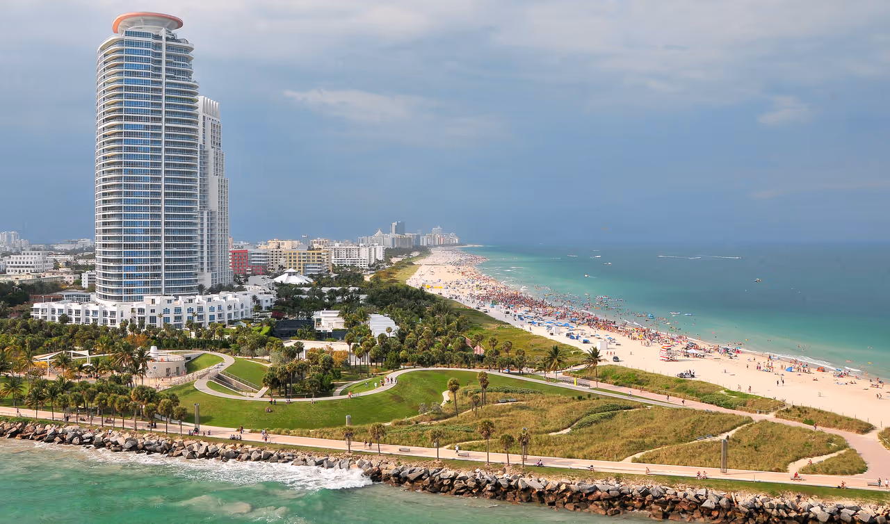 Aerial view of a coastal park and sandy beach in South Beach, Miami, with a prominent skyscraper under a cloudy sky