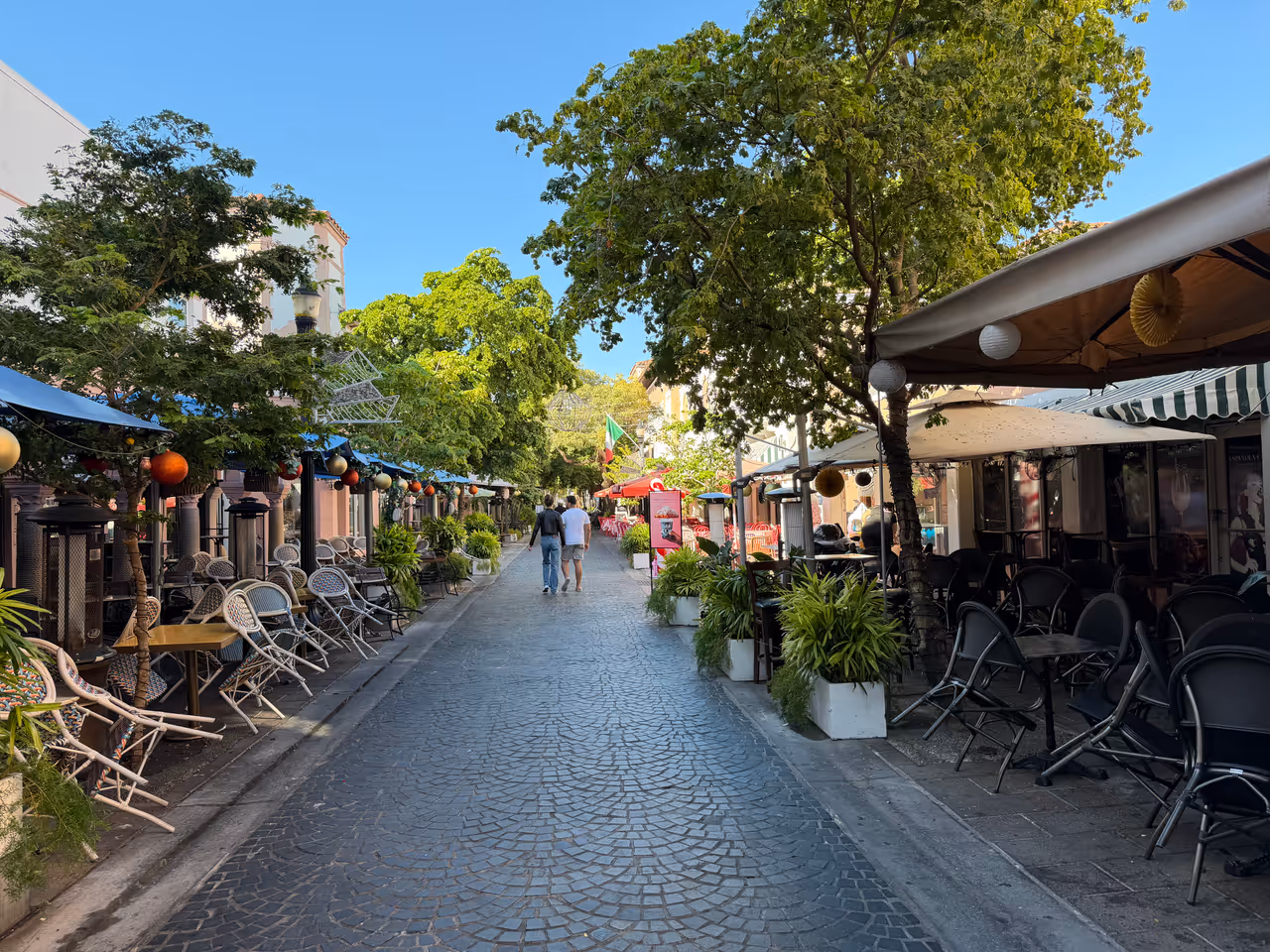 Pedestrianised cobblestone street of Espanola Way in South Beach, Miami, Florida, lined with outdoor restaurant seating, café tables under umbrellas and lush green trees
