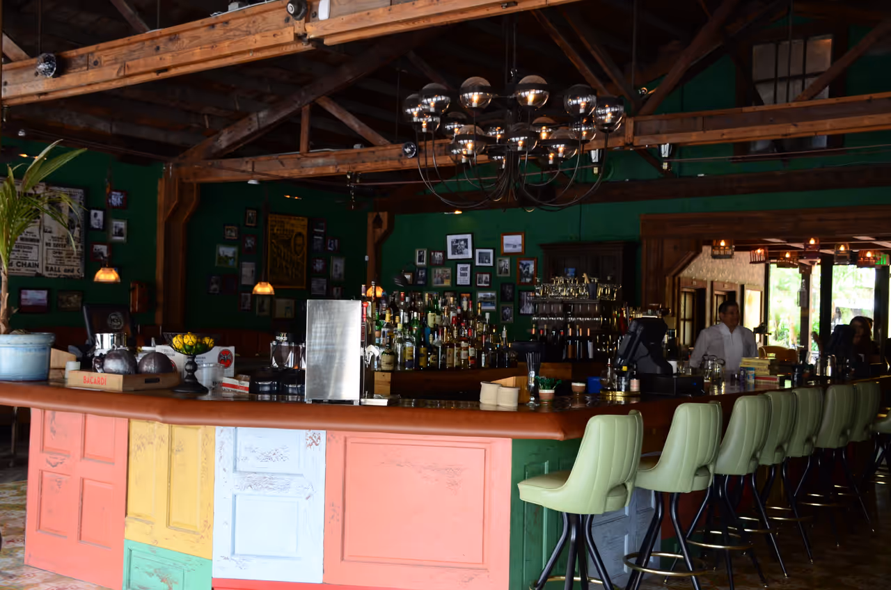 Inside of a vibrant bar in Little Havana, Miami, featuring a colorful panelled bar counter, light green bar stools, green walls, and exposed wooden beams